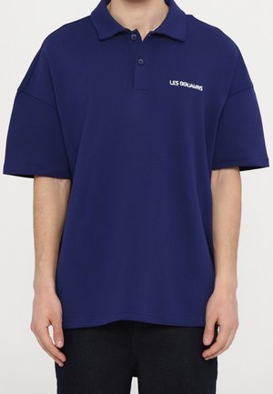 Polo azul marino de manga corta, con tapeta de dos botones y "LES BENJAMINS" bordado en el pecho izquierdo, llevado por una persona.