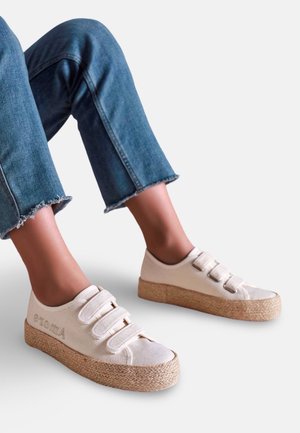 Personne portant des baskets en toile beige avec trois bandes auto-agrippantes et des semelles tissées épaisses, assorties à un jean bleu raccourci.