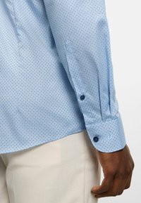 Hellblaues Hemd mit geometrischem Muster, langen Ärmeln und kontrastierenden blauen Knöpfen an der Manschette, in hellbeige Hosen gesteckt.