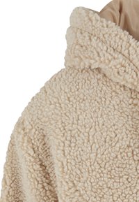 Detailný záber na béžovú sherpa flísovú tkaninu s hrubými, textúrovanými, kučeravými vlnenými vláknami a preloženým okrajom goliera.