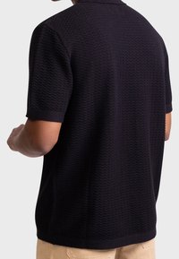 Musta teksturoidulla siksak-kuviolla varustettu poolopaita, jossa on lyhyet hihat ja resorikaulus, nähtynä takaa.