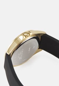 Montre dorée avec un bracelet en silicone noir. Dispose d'un boîtier rond, d'une couronne texturée et d'un dos en métal lisse. Aucune inscription visible.