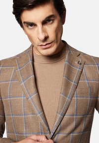 Brązowa marynarka w wzory z niebieskimi akcentami w kratę, wykonana z teksturowanej tkaniny, nałożona na brązowy sweter z golfem o prążkowanej strukturze. Zapięcie na podwójny guzik.