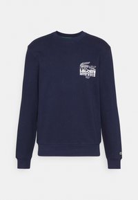 Felpa blu navy con maniche lunghe, colletto rotondo, caratterizzata da un logo Lacoste bianco e un grafico di coccodrillo sul lato sinistro del petto.
