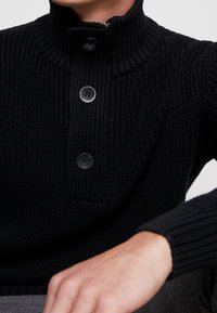 Czarny sweter dziany z wysokim prążkowanym kołnierzem, z trzema dużymi guzikami i fakturowanym wzorem ściegu.