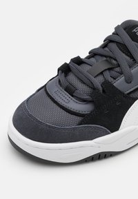 Chaussure de sport grise en mélange de matériaux en maille et en daim. Présente une languette rembourrée, un design superposé et une semelle en caoutchouc blanc.