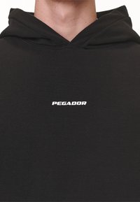 Sudadera negra con capucha ajustable y un logo blanco "PEGADOR" en el frente. Textura de tela suave, corte relajado y diseño minimalista.