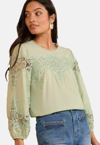 Blusa verde chiaro con dettagli in pizzo intricati sulle maniche lunghe e sul davanti, scollo rotondo e morbida texture del tessuto; abbinata a jeans a vita alta.