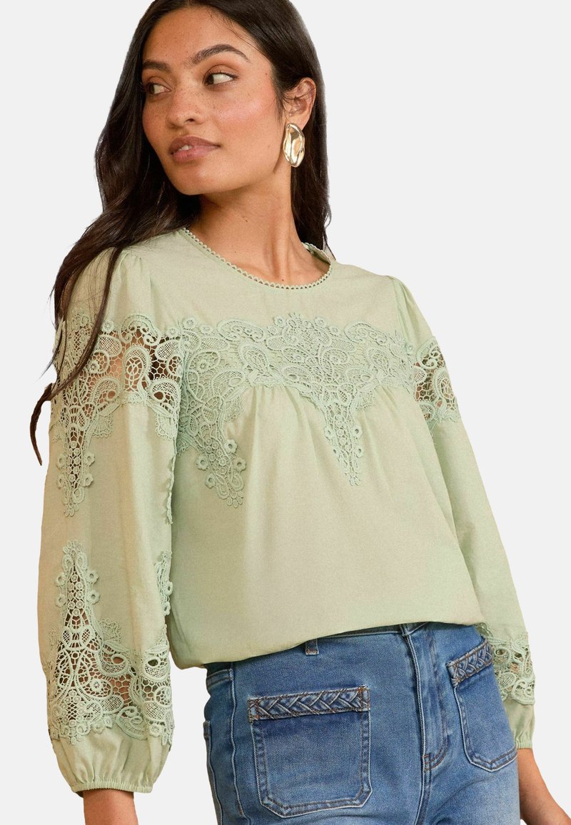 Blusa verde chiaro con dettagli in pizzo intricati sulle maniche lunghe e sul davanti, scollo rotondo e morbida texture del tessuto; abbinata a jeans a vita alta.