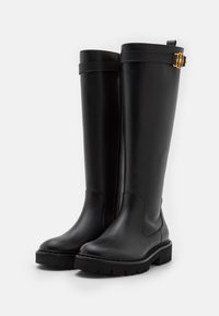Bottes imperméables noires montant jusqu'aux genoux en cuir lisse. Elles présentent une semelle à relief texturé et un accent en métal doré au sommet.