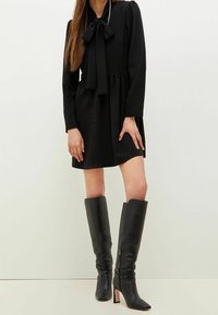 Frau in einem schwarzen, langärmligen Kleid mit einer Schleifen-Nekkerl, kombiniert mit knielangen, schwarzen Lederstiefeln mit Absatz, stehend vor einem einfarbigen Hintergrund.