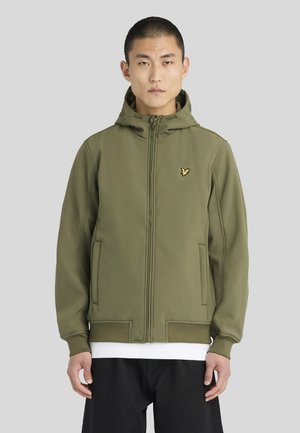 Homme portant une veste à capuche zippée verte olive avec un petit logo oiseau jaune sur la poitrine, debout devant un fond gris uni.