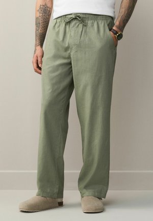 Férfi világos zöld, húzózsinóros nadrágban, bézs papucsban, fehér ingben, kar tetoválásokkal és jobb csuklóján órával.