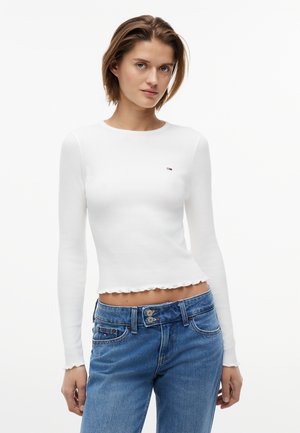 Žena v bielom krátkom top s dlhým rukávom a malým logom na hrudi, v modrých rifle s vysokým pásom, na jednoliatom pozadí.