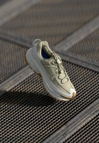 Sapatilha atlética de beige claro com detalhes castanhos e sola grossa, exposta numa superfície de grelha metálica sob luz natural.