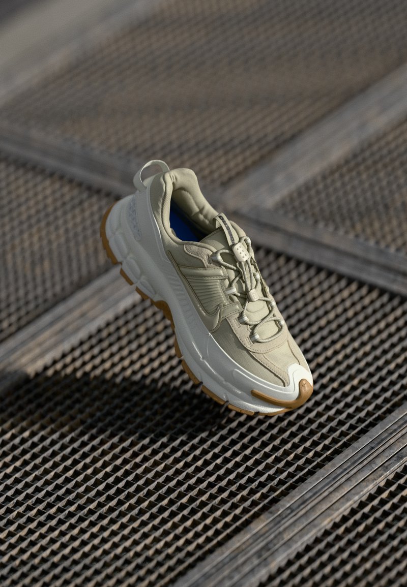 Sapatilha atlética de beige claro com detalhes castanhos e sola grossa, exposta numa superfície de grelha metálica sob luz natural.