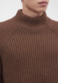 Jeune personne portant un pull en tricot côtelé marron avec col montant, visible du cou jusqu'à la poitrine sur un fond uni.