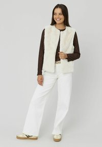 Gilet en fausse fourrure blanche avec un col arrondi, associé à un haut en dentelle noir à manches longues et à un jean blanc à jambes larges. Des chaussures décontractées complètent le look.