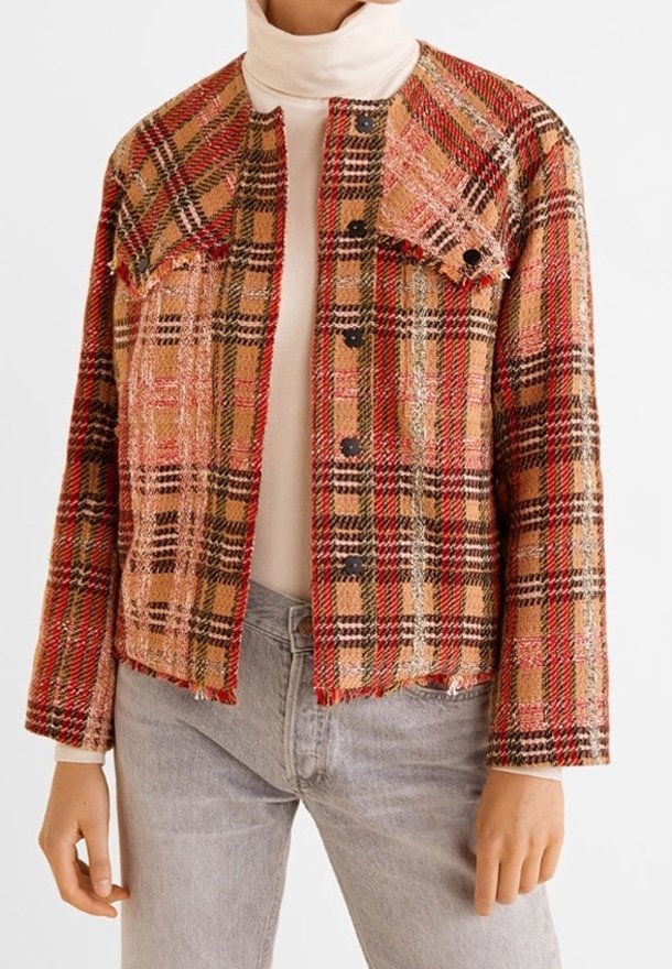 Une femme porte une veste courte à carreaux aux tons rouge, beige et noir par-dessus un col roulé blanc et un jean gris clair.