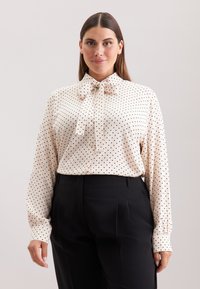 Crèmeblouse met zwarte stippen, lange mouwen en een strik bij de hals; gecombineerd met zwarte hoge taillebroeken. Gladde textuur.