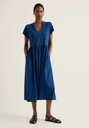 Vrouw staat met handen in zakken, draagt een blauwe midi-jurk met V-hals en zwarte platte sandalen tegen een lichte achtergrond.