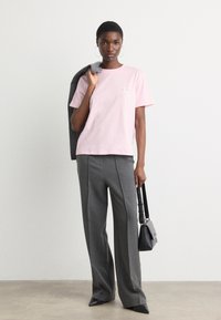 Light pink cotton t-shirt with short sleeves, paired with gray high-waisted trousers. Model holds a gray jacket and black bag.
