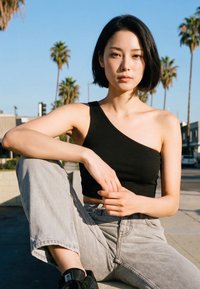 Giovane donna con top nero a una spalla e jeans grigi seduta all'aperto in una giornata di sole con palme e edifici sullo sfondo.