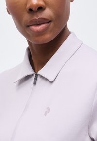 Κοντινό ενός ατόμου που φορά ανοιχτογκρι πουκάμισο με φερμουάρ και γιακά, εστιάζοντας στην κάτω περιοχή του προσώπου και στο πάνω μέρος του στήθους.