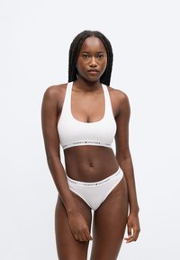 Valge spordirinnahoidja ja sobivad aluspüksid logoga. Rinnahoidjal on ümardatud kaelus ja laiad rihmad. Sile, puuvillale sarnane tekstuur.