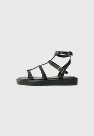 Black leather sandal with flat sole, featuring three straps across the foot and an adjustable ankle strap with a gold buckle.