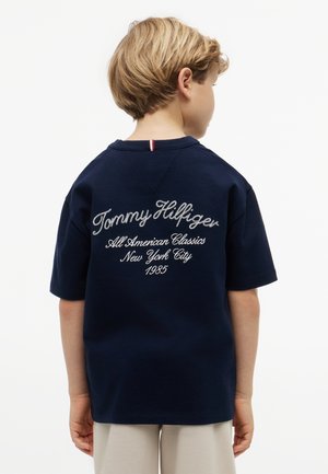 Barn med blondt hår iført en mørkeblå Tommy Hilfiger T-shirt med teksten "All American Classics New York City 1985" på ryggen.