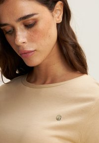 Camiseta beige con cuello redondo, textura suave y pequeño detalle de logo metálico en el lado izquierdo. Diseño suave y ajustado.