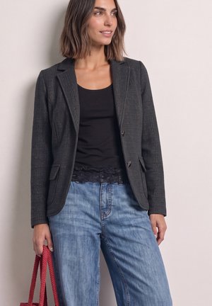 Donna con capelli fino alle spalle che indossa un blazer scuro, un top nero, jeans blu, tiene una borsa intrecciata rossa e guarda di lato.