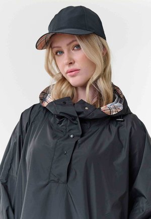 Donna con capelli biondi indossa un berretto nero e un impermeabile nero con fodera a quadri beige sul colletto, su uno sfondo chiaro uniforme.