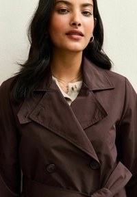 Trench coat marrone doppiopetto con vita aderente, ampi revers e dettagli con bottoni, indossato sopra un top chiaro e collane sovrapposte.