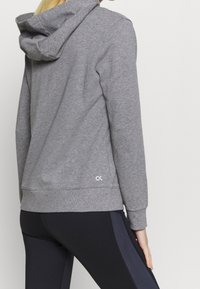 Een persoon in een grijze hoodie en zwarte leggings met donkere zijstrepen, gefotografeerd van achteren tegen een effen achtergrond.