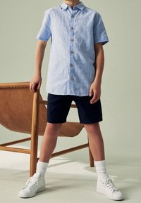 Enfant debout à côté d'une chaise en bois, portant une chemise à manches courtes bleu clair boutonnée, un short marine, des chaussettes blanches et des baskets blanches.
