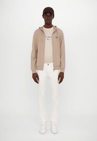 Chaqueta beige con cremallera y capucha, camiseta crema y pantalones blancos. Zapatillas blancas completan el look casual. Diseño sencillo con acentos mínimos.