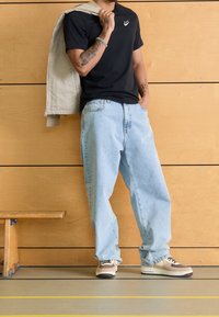 Black short-sleeve shirt with white logo, paired with light blue baggy jeans and brown sneakers with white accents. Benchtop and wood backdrop.