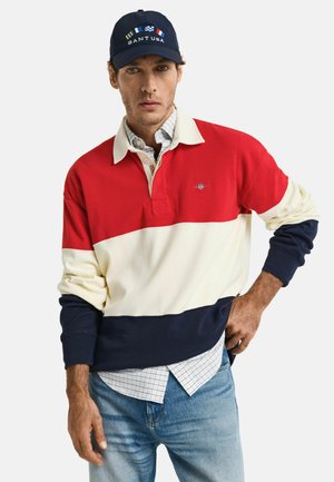 Rood, wit en marineblauw gestreepte rugby-stijl pullover met een kraag, contrasterende mouwen en geborduurd logo; gedragen over een geruite shirt.