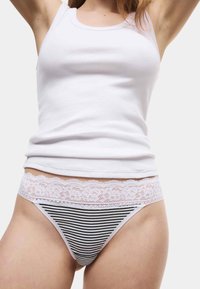 Frau, die ein weißes Tanktop und schwarz-weiß gestreifte Unterwäsche mit Spitzenbesatz trägt, mit erhobenen Armen vor einem schlichten Hintergrund.