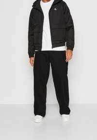 Svart zip-up jacka med huva och framfickor, tillsammans med en vit T-shirt och svarta vida byxor, kompletterat med vita sneakers.