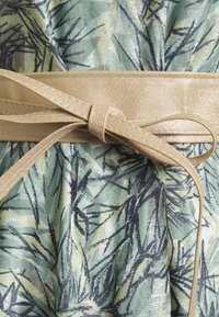 Glittrande beige bälte knytet i en rosett runt ett tyg med grön, blå och beige abstrakt bladmönster.