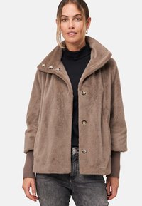 Veste en fausse fourrure beige avec un large col, dotée de boutons en argent et de poignets côtelés. Le design est ample avec une texture douce.