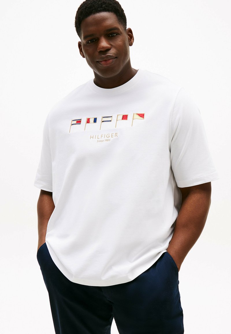 Biała bawełniana koszulka z haftowanymi flagami morskimi oraz napisem „HILFIGER”. Okrągły dekolt i krótkie rękawy. Luźny krój, minimalistyczny design.