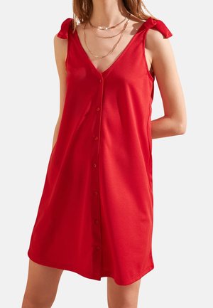 Femme portant une robe rouge sans manches à boutons, avec des bretelles nouées aux épaules et des colliers en chaînes d'or superposés.