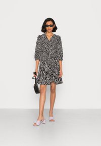 Vestido preto envolvente com padrão de bolinhas brancas, mangas três quartos e detalhe franzido na lateral. Combinado com sandálias block heel lavanda e uma bolsa preta.