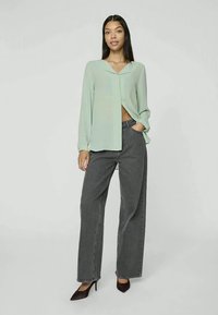 Lys grønn bluse med åpen front og lange ermer, kombinert med høy midje grå vide jeans. Modellen står i spisse hæler.