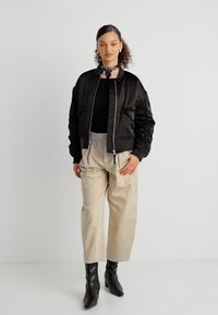 Γυναίκα φορώντας μαύρο μπουφάν τύπου μπόμερ, μαύρο τοπ, μπεζ παντελόνι ψηλόμεσο, μαύρα μποτάκια αστραγάλου και ένα μικρό λαιμοκούφι με μοτίβο.