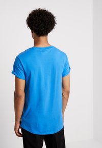 Blå bomullströja med uppvikt ärm, rund hals och rak fåll, med en jämn textur. Sedd bakifrån.